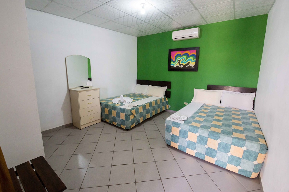 A hotel room with two beds, a dresser, a bench, a mirror, and a green wall with a colorful painting.