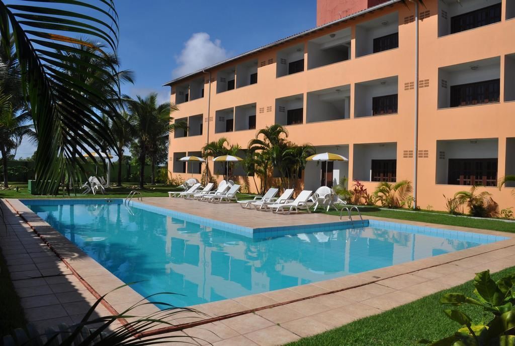 Una piscina con sillas de descanso y sombrillas, rodeada de palmeras y un edificio con varios balcones.