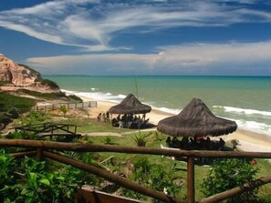 A beach view with thatched umbrellas, a wooden walkway, and a rocky cliff. - Quiriri Park Hotel (Prado)