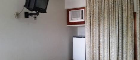 Une petite pièce avec un téléviseur fixé au mur, un réfrigérateur et une fenêtre avec des rideaux.