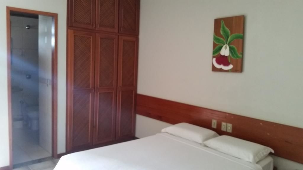 Quarto com cabeceira de madeira, cama com roupa de cama branca, guarda-roupa de madeira e um quadro de uma flor na parede.