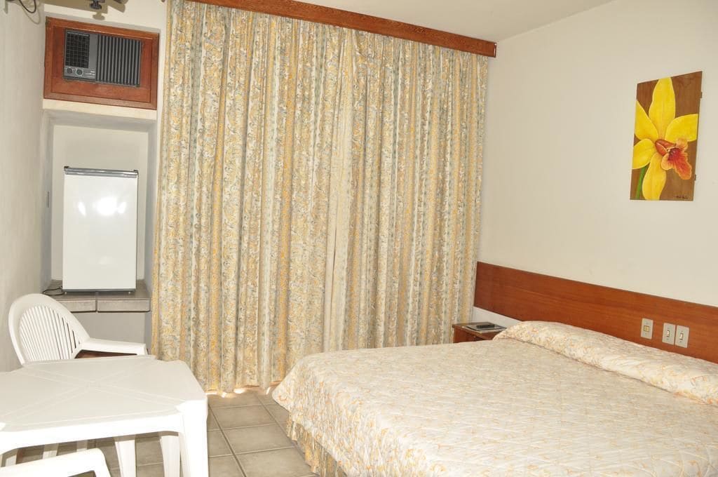 A hotel room with a bed, a desk, a chair, a whiteboard, and a painting of a flower on the wall.