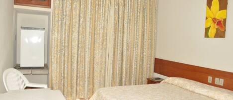 A hotel room with a bed, a desk, a chair, a whiteboard, and a painting of a flower on the wall.
