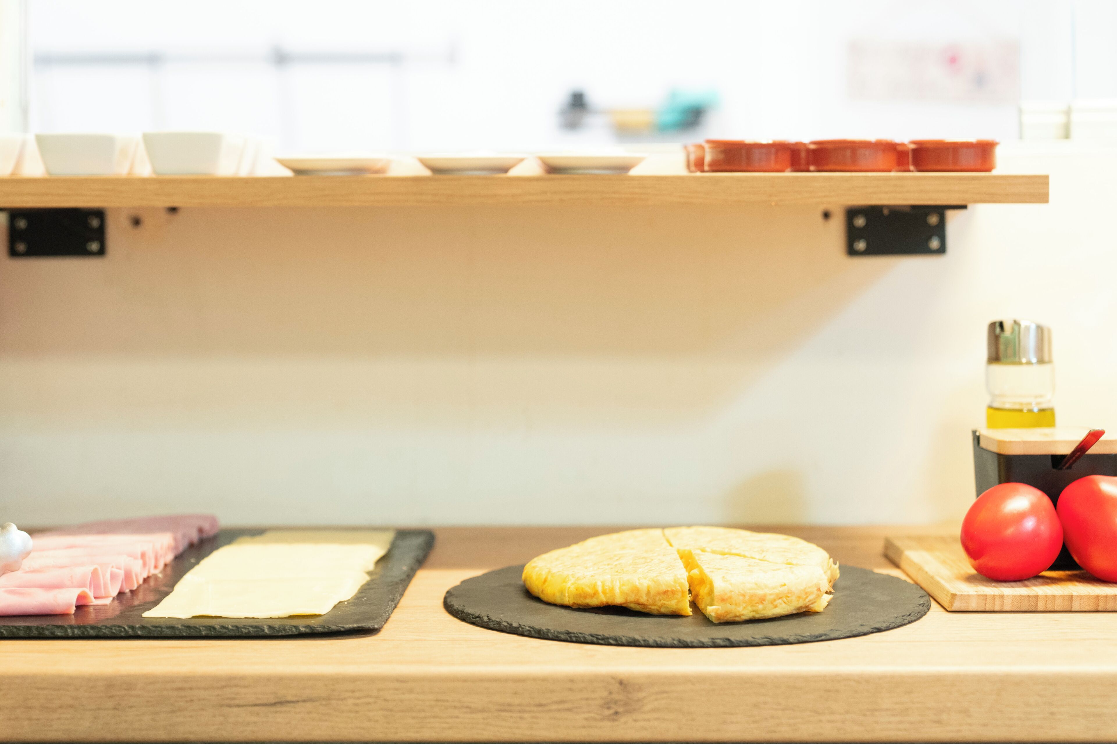 A kitchen counter with a rolled omelette on a black plate, a cutting board with sliced ham, and a wooden shelf above holding various kitchen items.