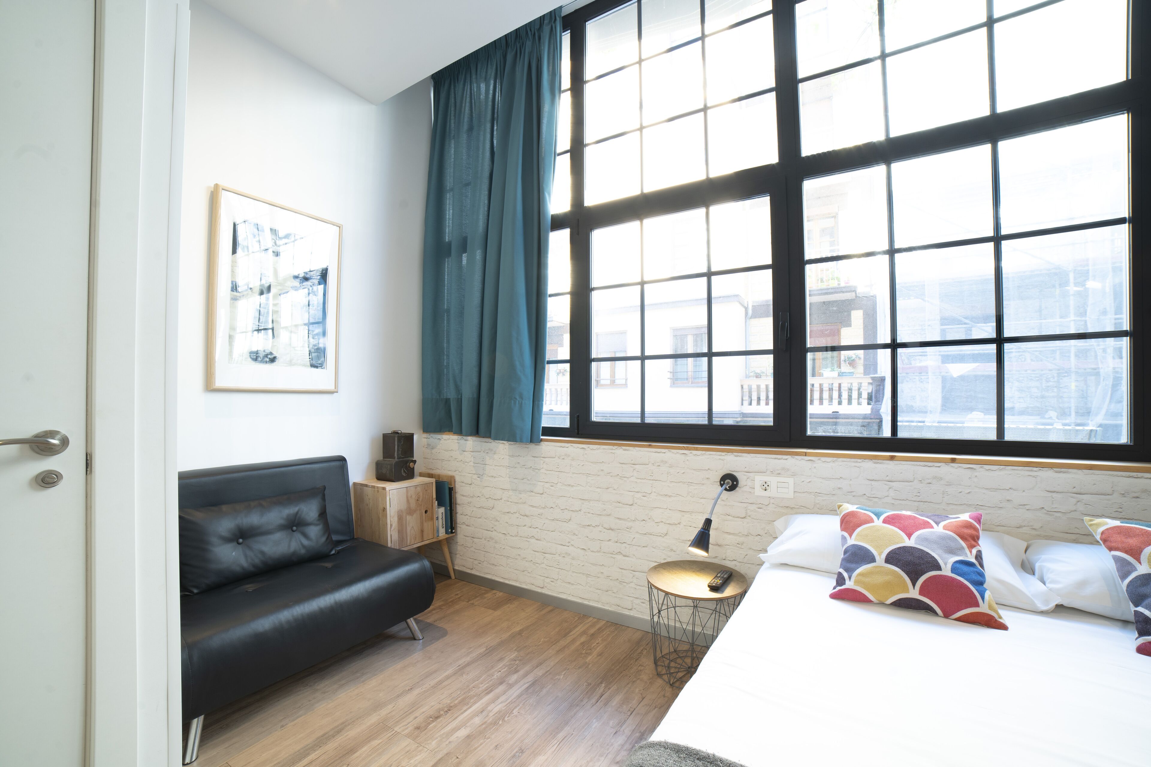 A modern room with a black leather sofa, a bed with colorful pillows, a small wooden side table, and a large window with a view of buildings.