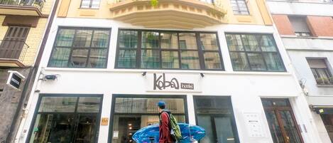 A building with a sign reading "Koba hostel" and a person carrying a blue surfboard.