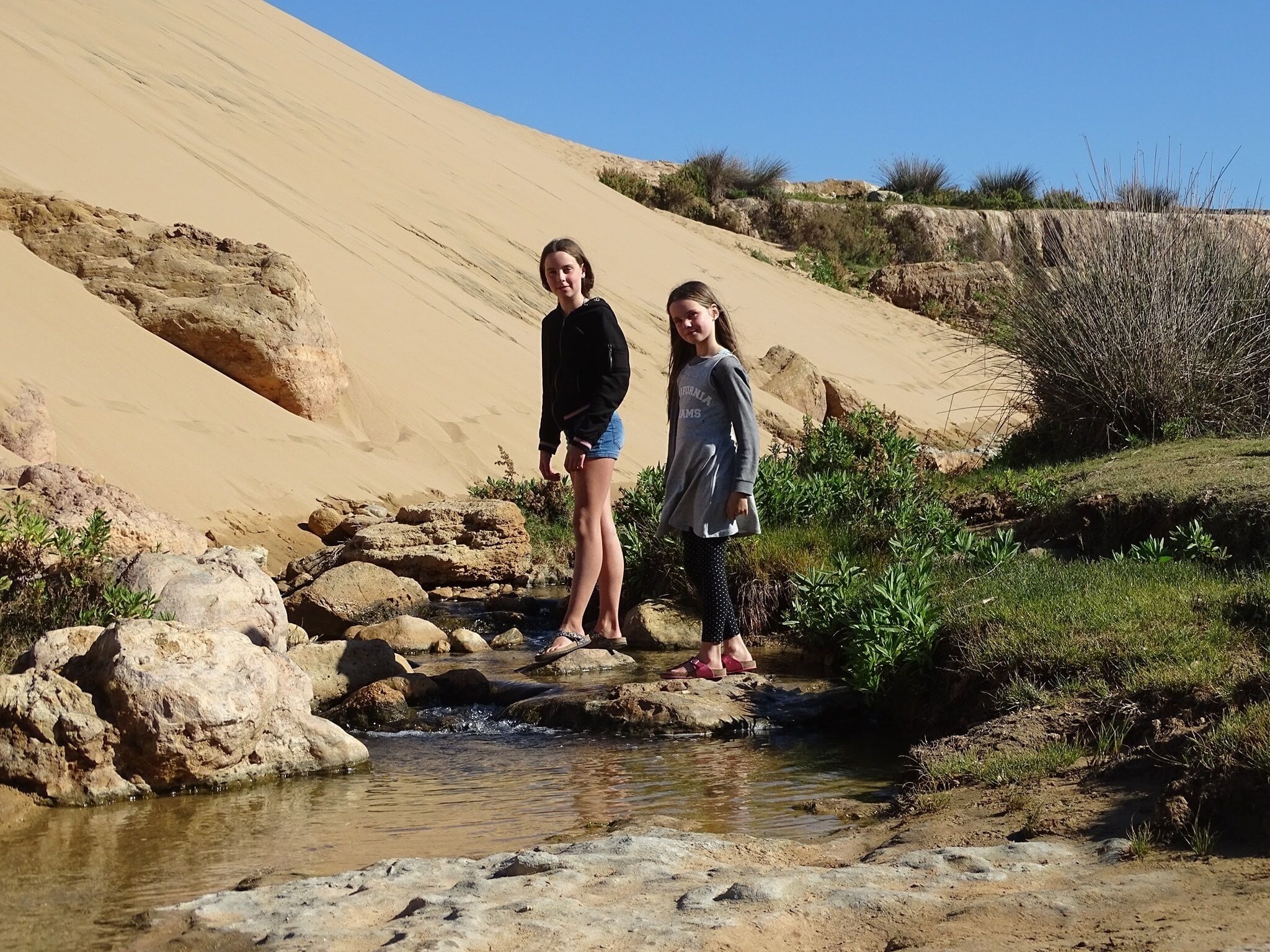Duas pessoas perto de um riacho em uma paisagem desértica, com dunas de areia ao fundo.