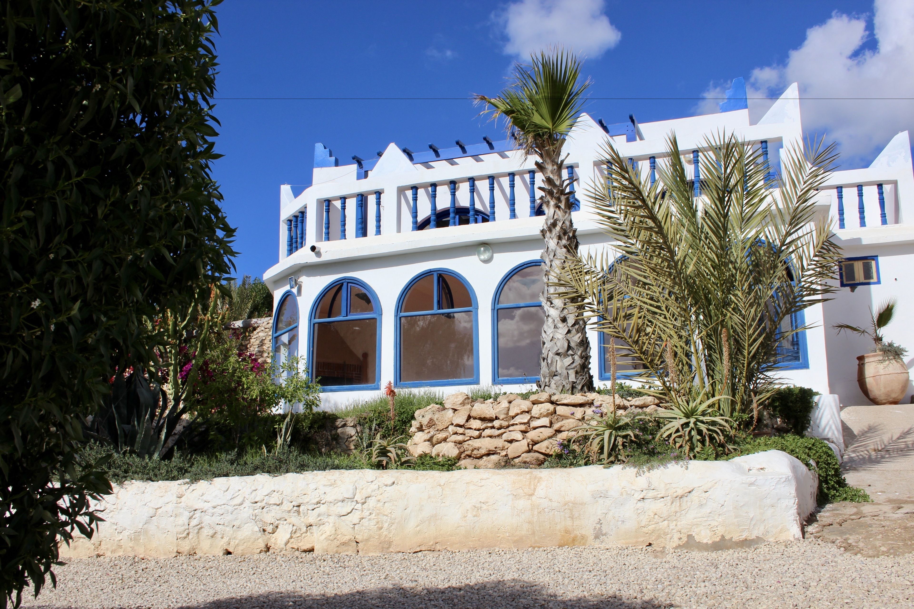 Un edificio blanco con detalles azules, ventanas arqueadas y una pared de piedra en primer plano.