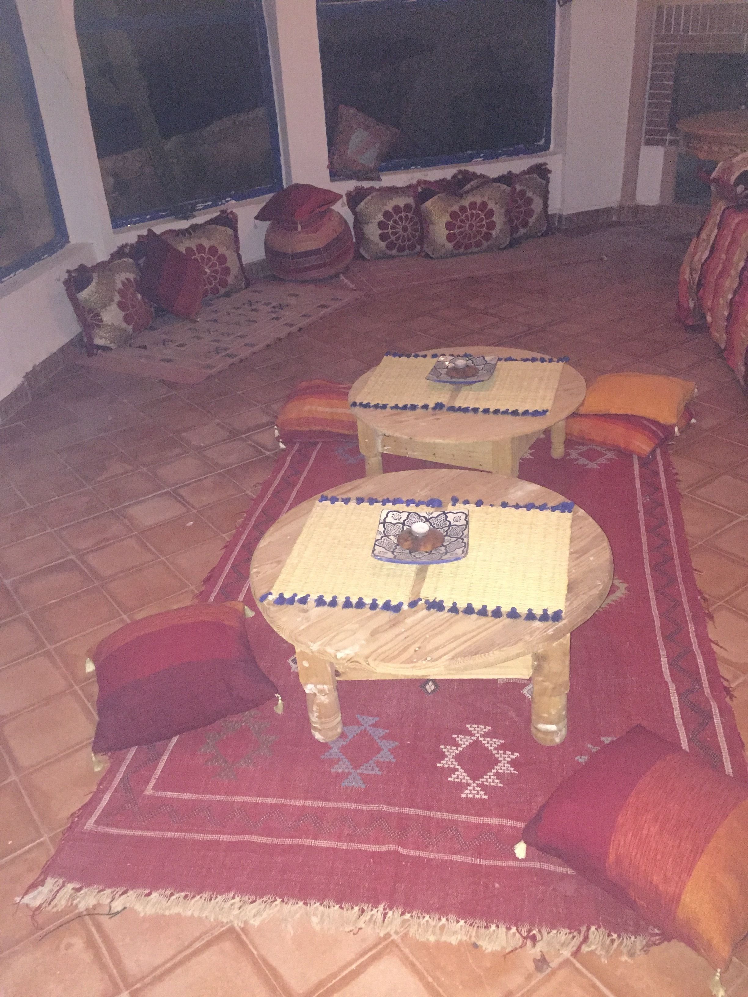 A room with two wooden tables, red and orange cushions, and a patterned rug.