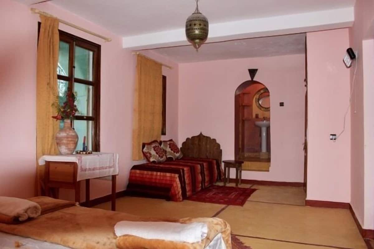 A room with a bed, a small table with a vase, a window with curtains, and a door leading to another room.
