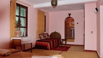 Una habitación con una cama, una mesita con un florero, una ventana con cortinas y una puerta que da a otra habitación.