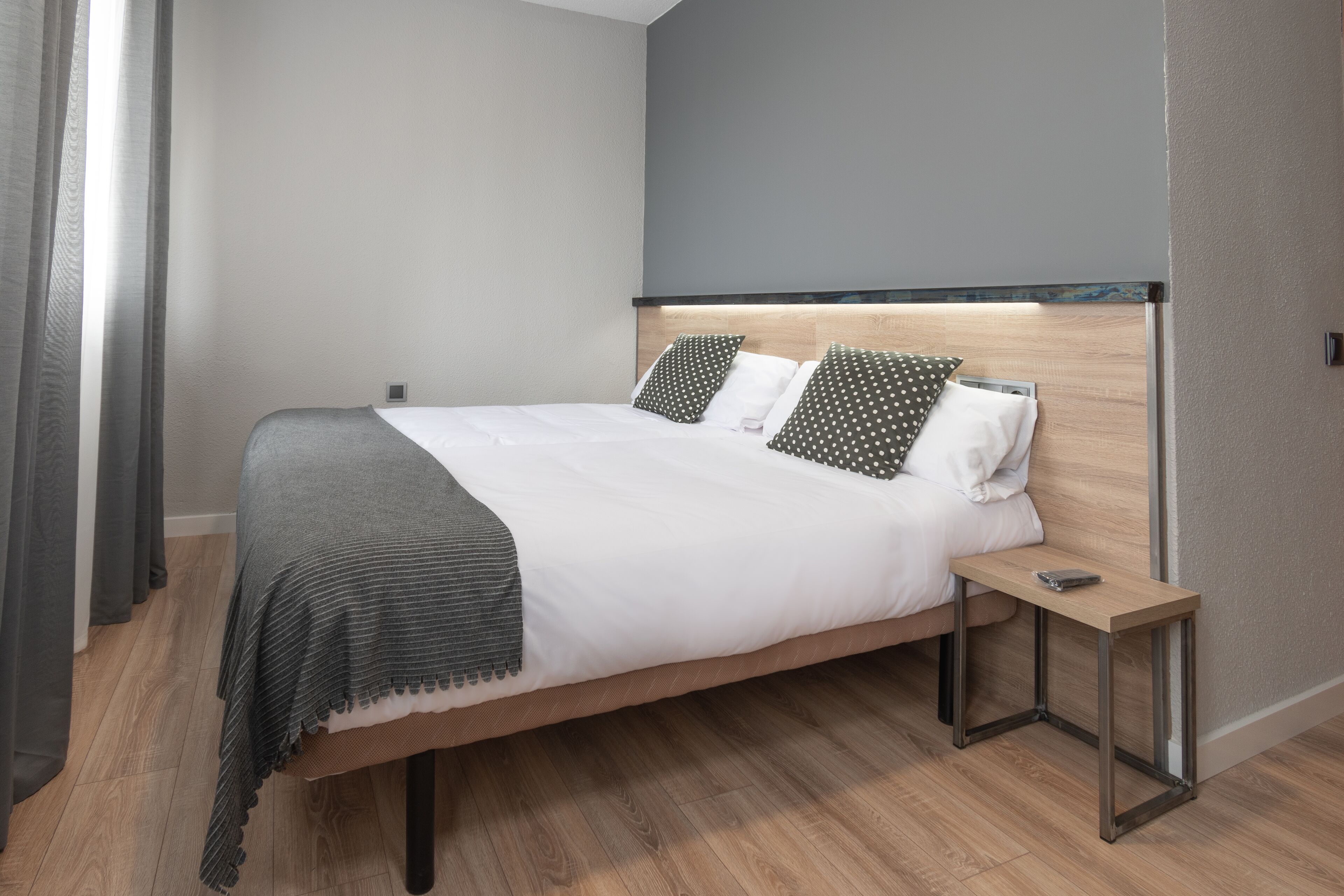 A modern hotel room with a wooden bed, grey curtains, and a small bedside table.