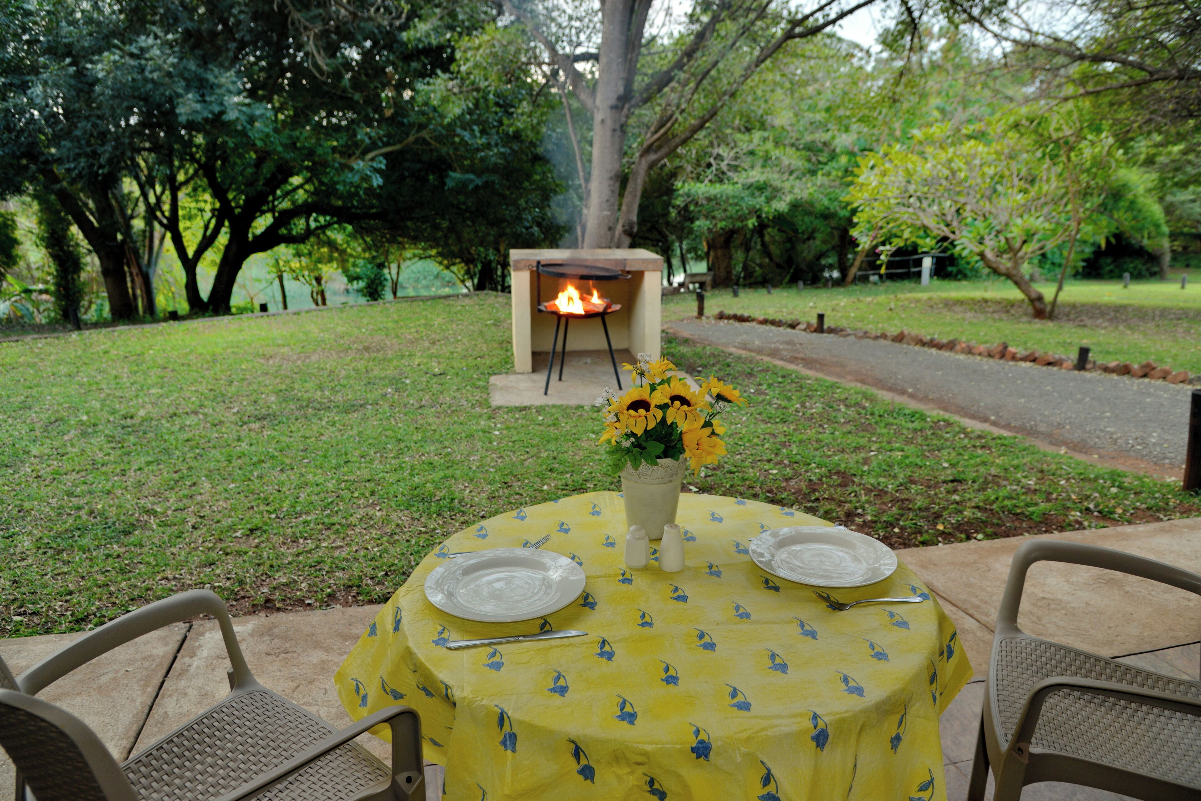 An outdoor dining area with a yellow tablecloth, two plates, a vase with flowers, and a wood-fired oven in the background.