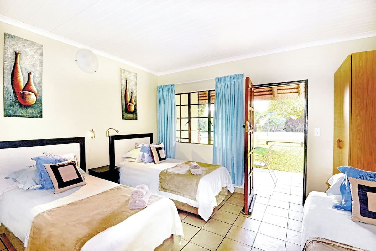 A hotel room with two beds, a large window with curtains, a view of the garden, and artwork on the walls.