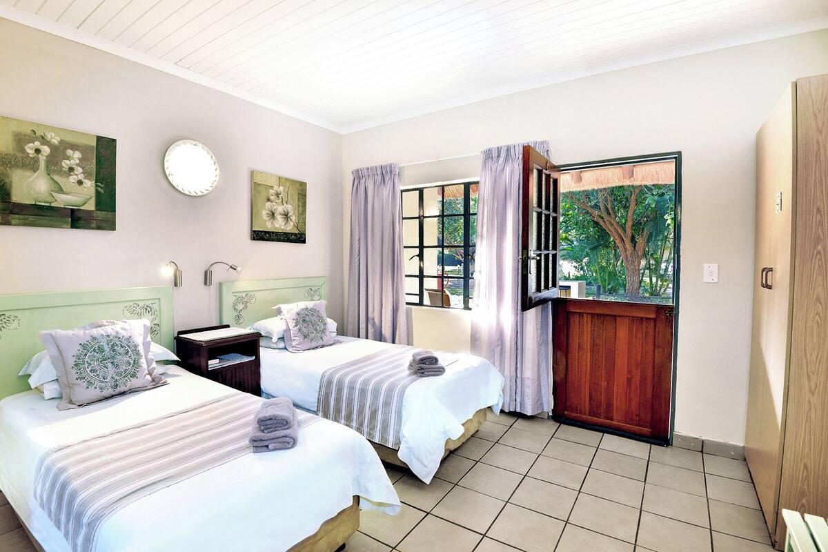 A bedroom with two beds, a window with curtains, a round mirror, and artwork on the walls.