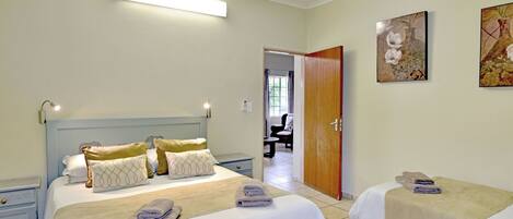 Une chambre d’hôtel avec un grand lit, un plus petit lit, un bureau et une chaise. Une porte donne accès à une autre pièce.