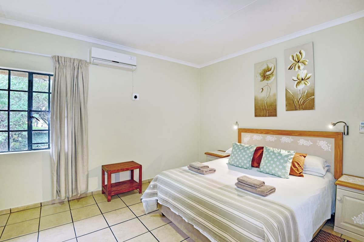 A hotel room with a large bed, a small bedside table, a nightstand, a window with curtains, and two wall-mounted flower paintings.