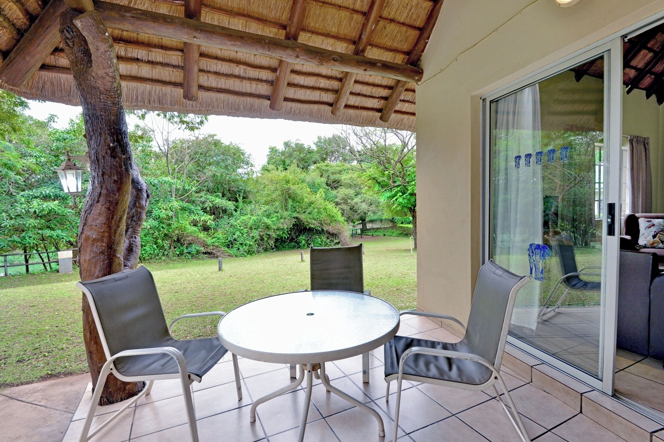 Un patio cubierto con una mesa redonda y sillas, una puerta de cristal que da a una habitación y vistas a un jardín exuberante.