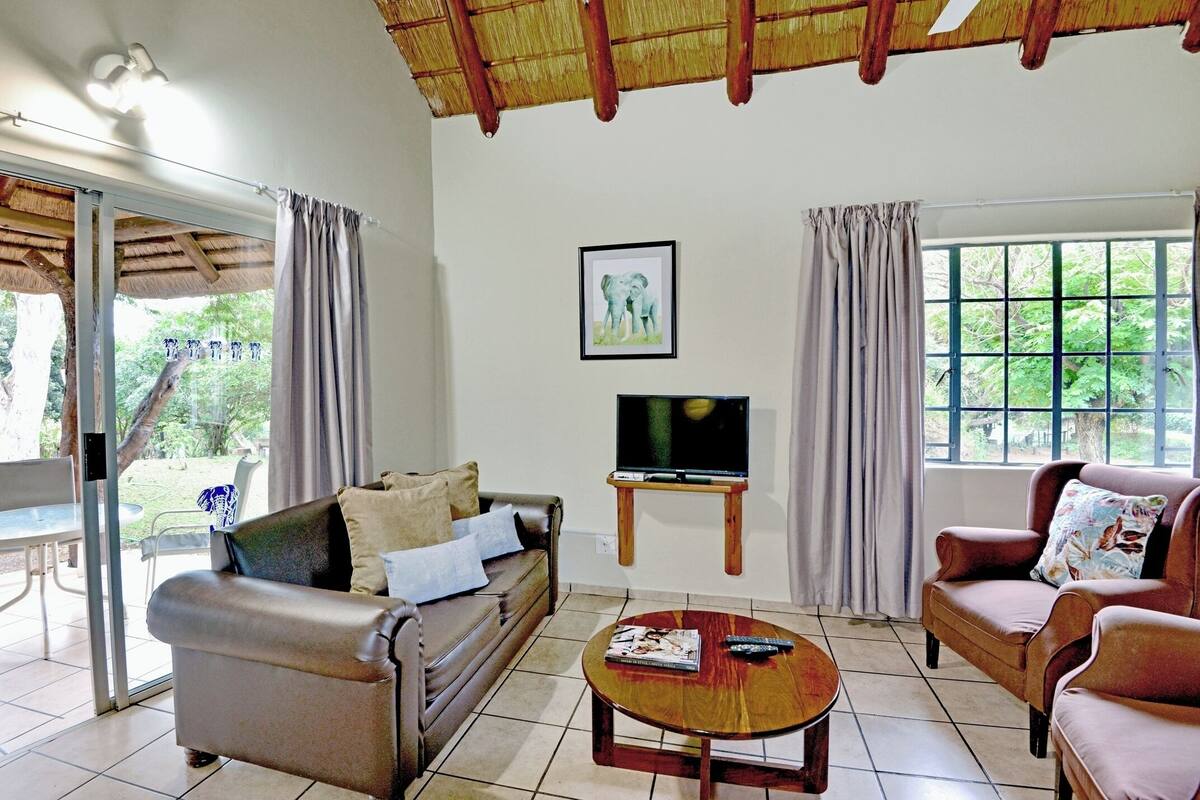 A living room with a sofa, armchair, and coffee table, a TV on a stand, and a window with curtains.