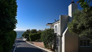 Una calle residencial con una casa que tiene balcón, vista despejada al océano y un jardín cuidado.