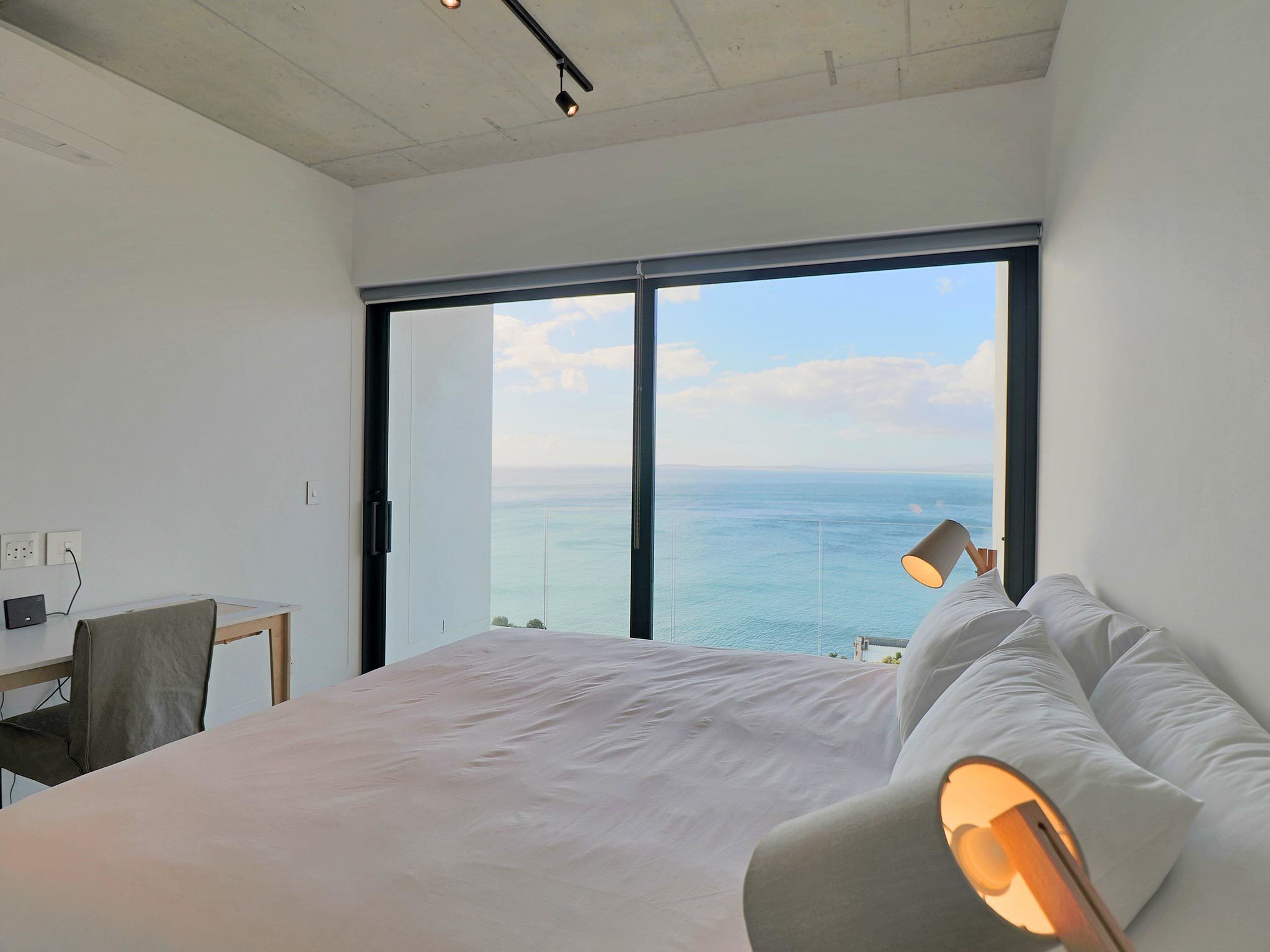 A bedroom with a large bed, a desk, and a view of the sea through a large window.