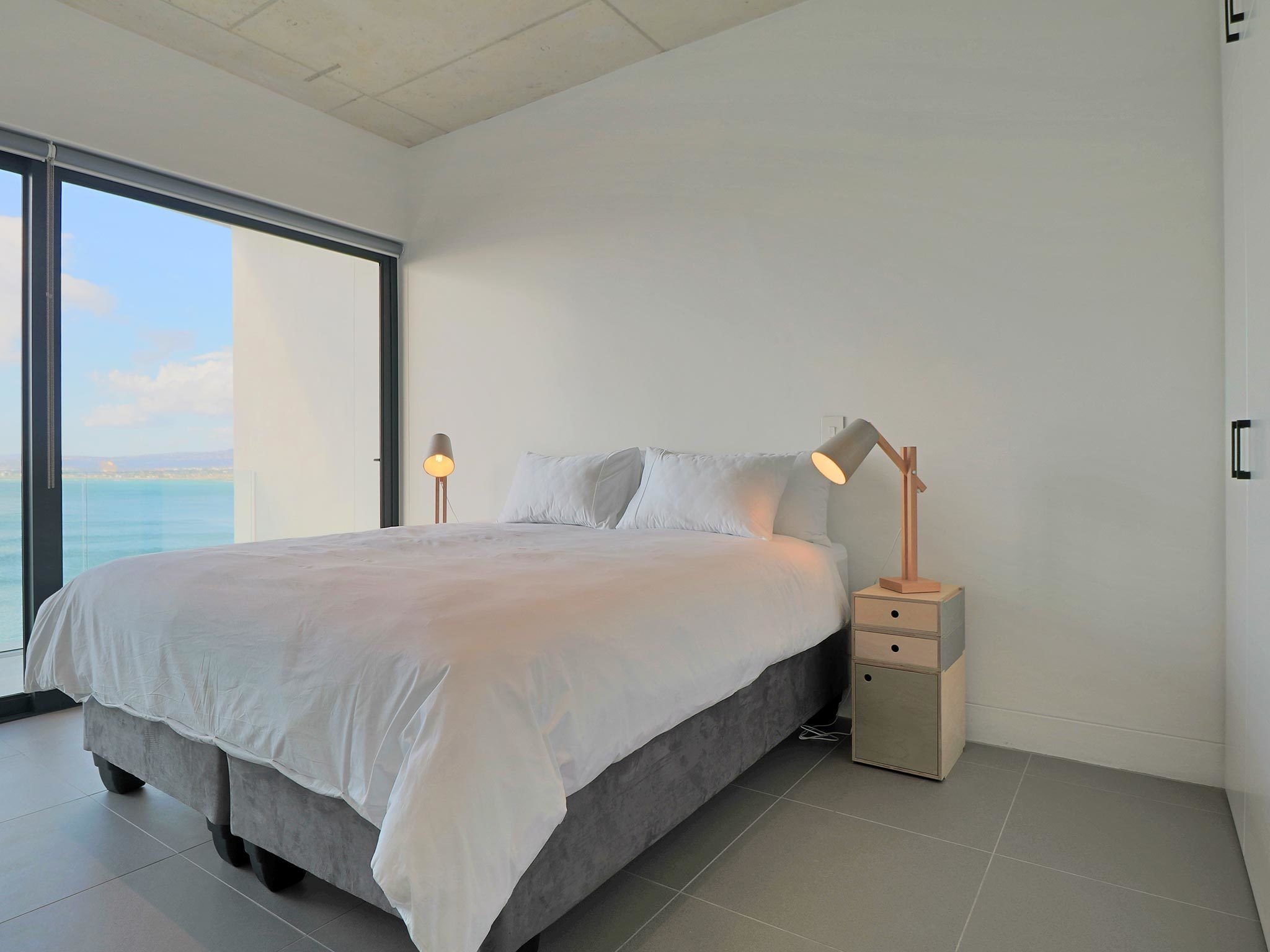 A modern bedroom with a large bed, two bedside tables, and a view of the sea through a large window.