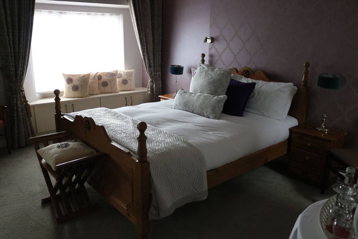 A neatly arranged bedroom with a wooden bed, a bedside table, a nightstand, a window with curtains, and a view of the outside.