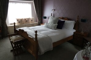 A neatly arranged bedroom with a wooden bed, a bedside table, a nightstand, a window with curtains, and a view of the outside. - New Park Hotel Athenry (Athenry)