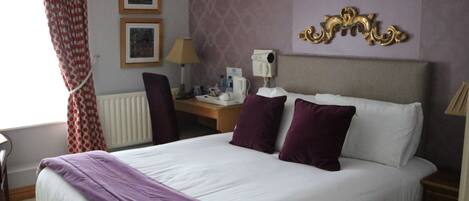 A hotel room with a large bed, a desk with a lamp, two framed pictures on the wall, and a radiator.