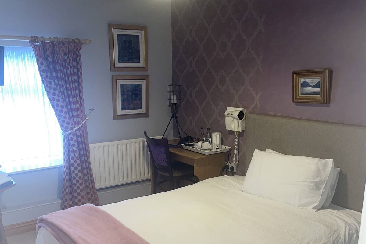 A bedroom with a bed, a desk, a chair, a radiator, and a window with curtains.