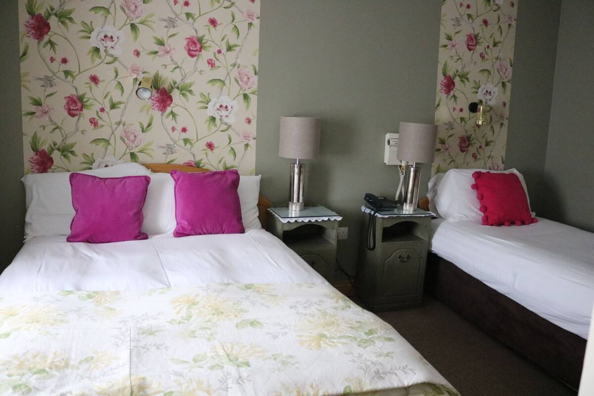 A hotel room with two beds, each with white bedding and pink pillows. There are two bedside tables with lamps and a phone. The walls have floral wallpaper.