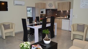 Un salon moderne avec un coin repas, comprenant un canapé, une table basse agrémentée d’une plante et de magazines, et un téléviseur fixé au mur.