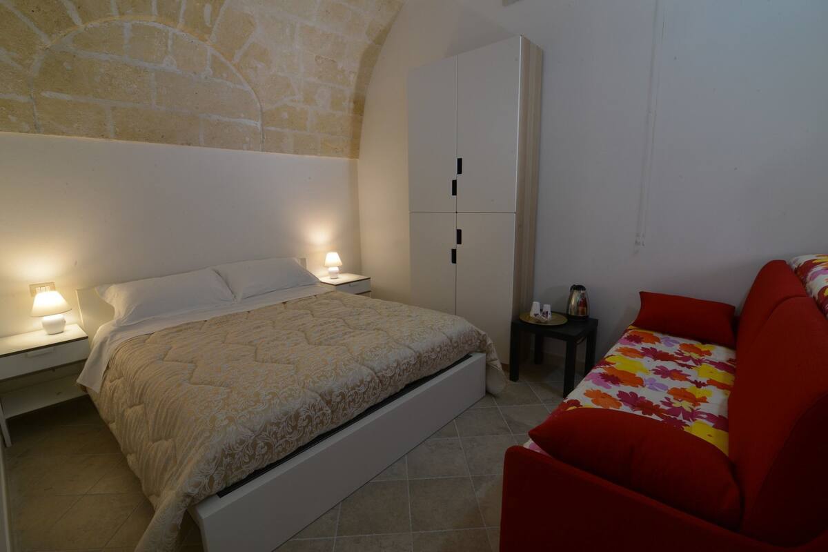 A bedroom with a bed, a wardrobe, a nightstand, a lamp, and a red sofa.