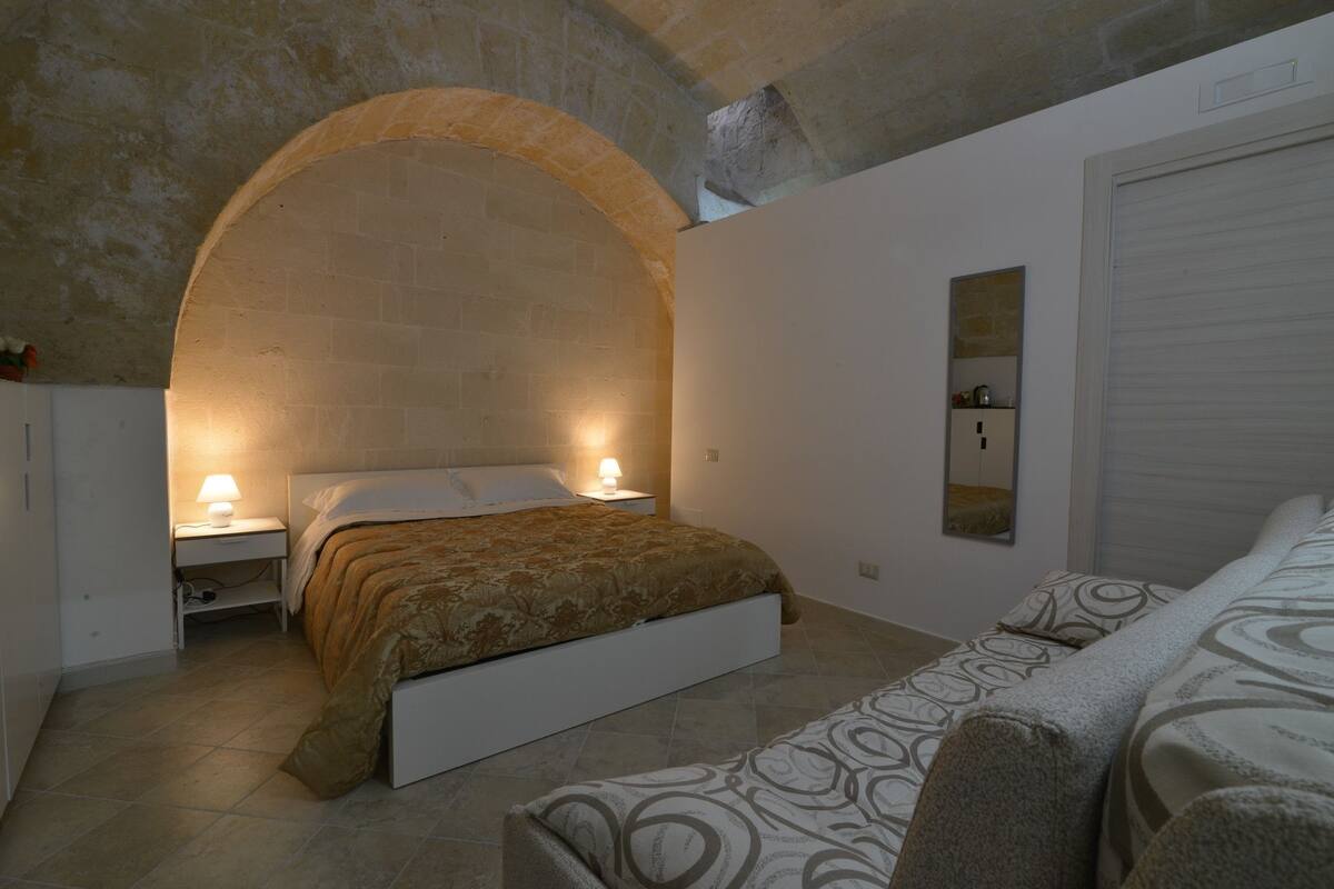 A bedroom with a bed, a nightstand, a lamp, a sofa, and a wall-mounted shelf.