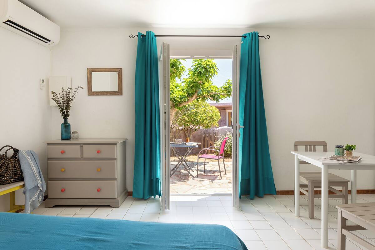A bedroom with a bed, a dresser, a desk, and a chair. There is a window with curtains, a mirror, and a vase with plants.
