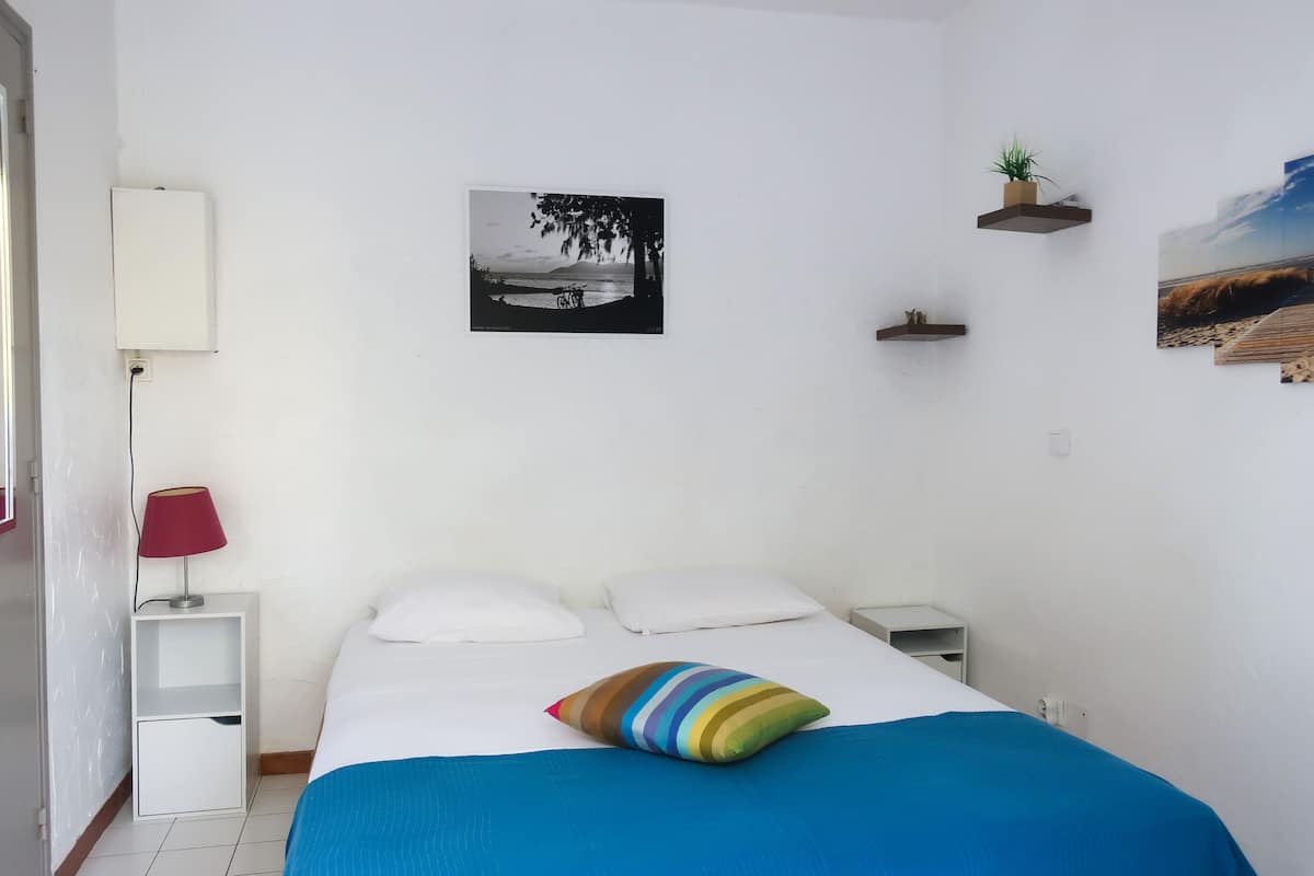 A neatly arranged bedroom with a bed, a bedside table with a lamp, a small shelf with a plant, and a wall-mounted picture.