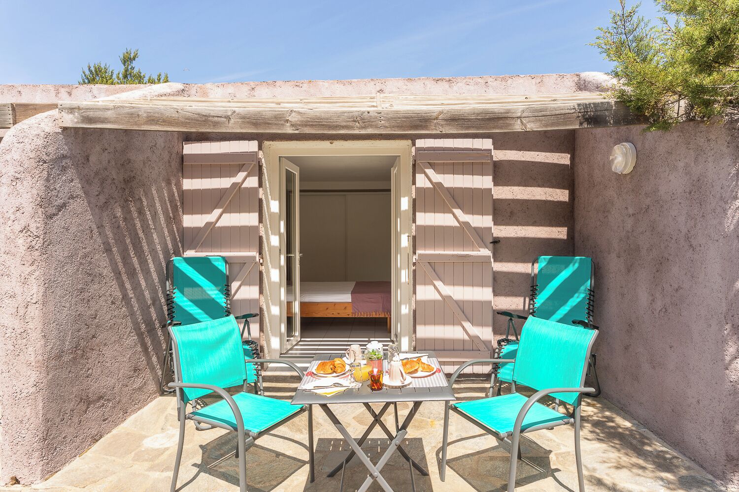 A small, enclosed outdoor space with a table and two chairs, a bed visible through an open door, and a concrete wall.