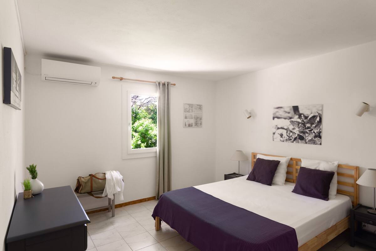 A bedroom with a bed, bedside tables, a desk, a chair, a window with curtains, and a wall-mounted air conditioner.