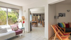 A spacious living area with a sofa, a dining table, and colorful chairs. A large window provides a view of greenery outside. - Domaine Bocca Di Feno (Bonifacio)