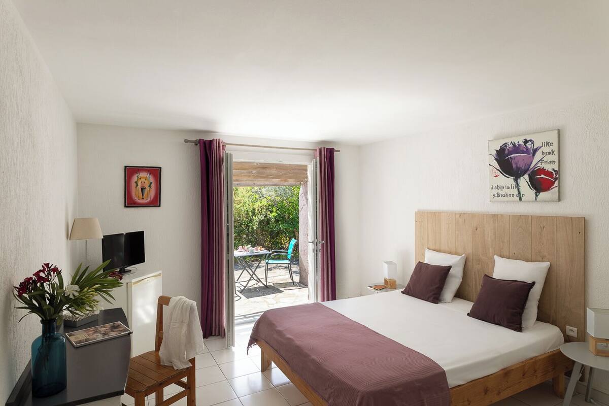 A bedroom with a bed, a desk with a computer, a chair, a small table with a lamp, and a window with curtains.
