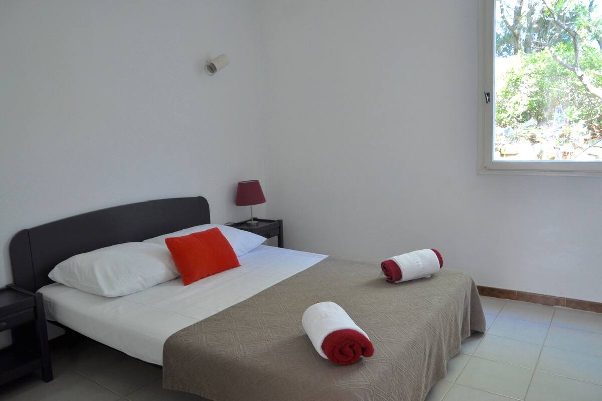 A neatly made bed with white and red pillows, a brown bedspread, and two rolled towels.