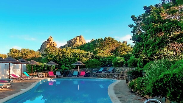 A pool area with lounge chairs and umbrellas, surrounded by greenery and a mountain backdrop. - Domaine Bocca Di Feno (Bonifacio)