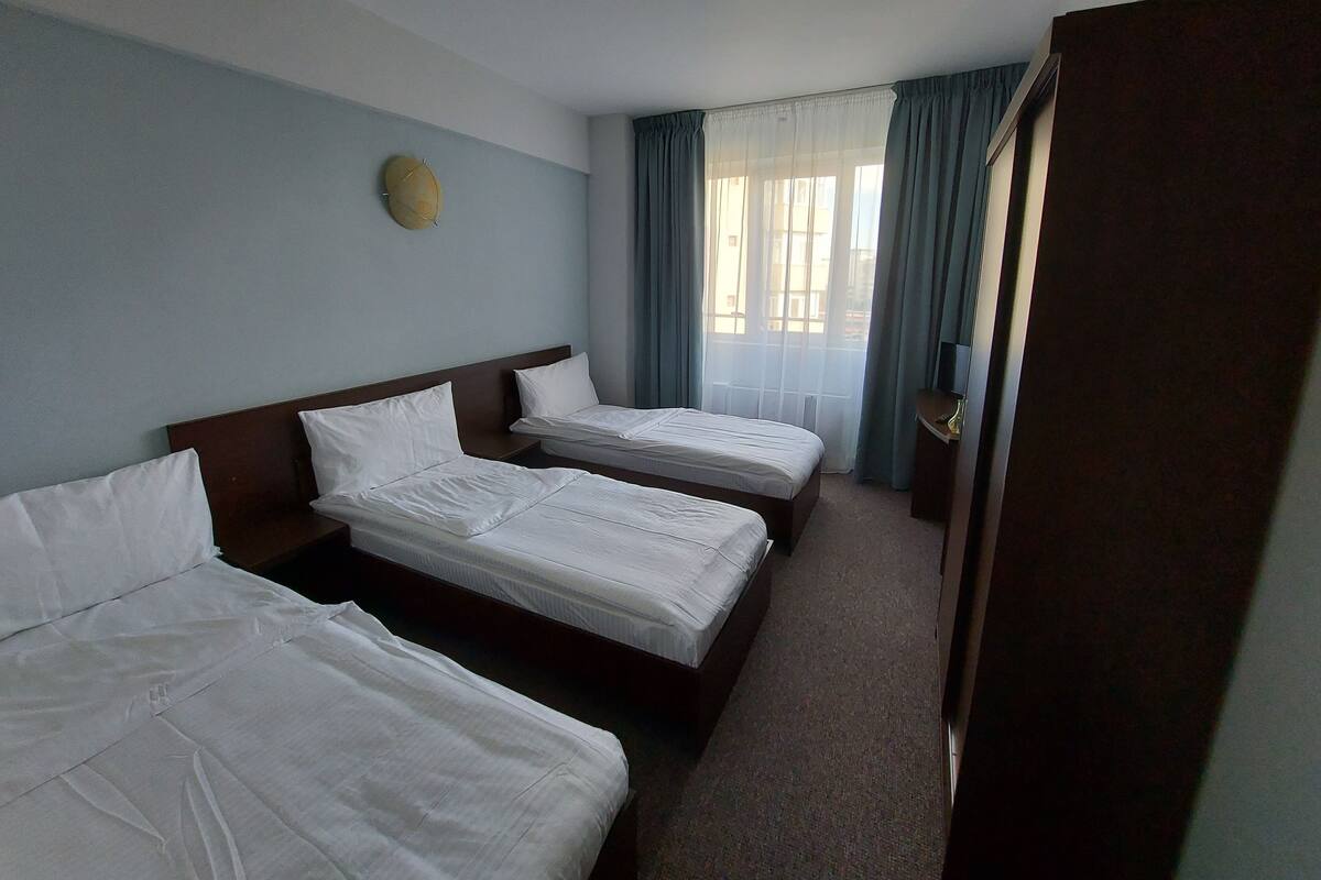 Habitación de hotel con dos camas individuales, cabecera de madera, un reloj redondo pequeño en la pared y una ventana con cortinas.