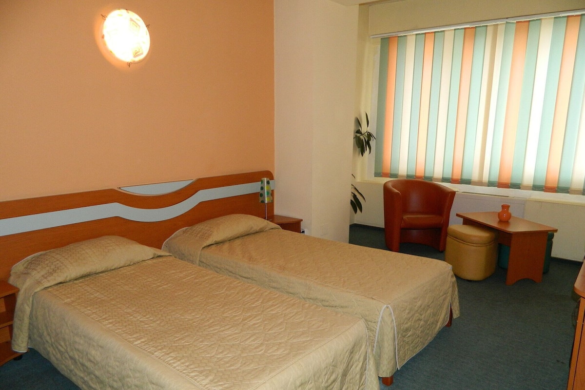 Habitación de hotel con dos camas, un escritorio, una silla y un ventanal con persianas a rayas.