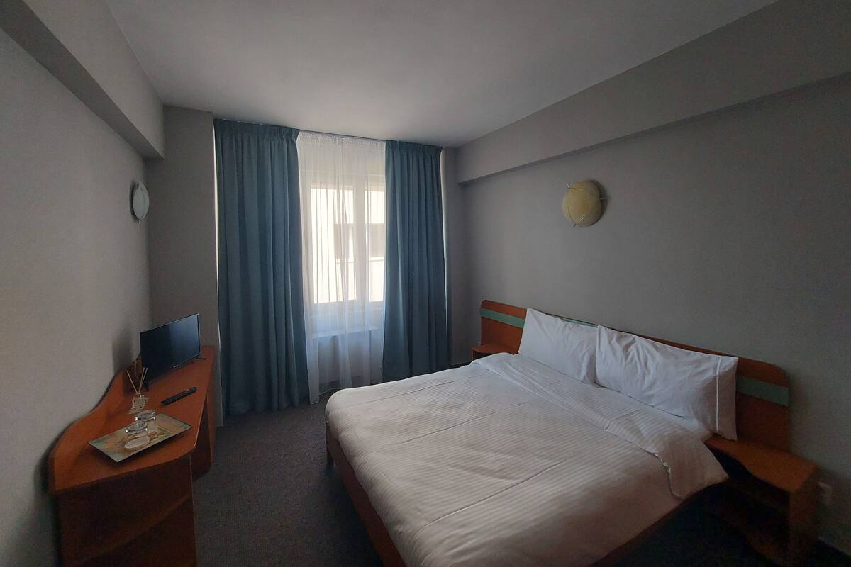 Una habitación de hotel con cama, escritorio, silla, reloj y ventana con cortinas.