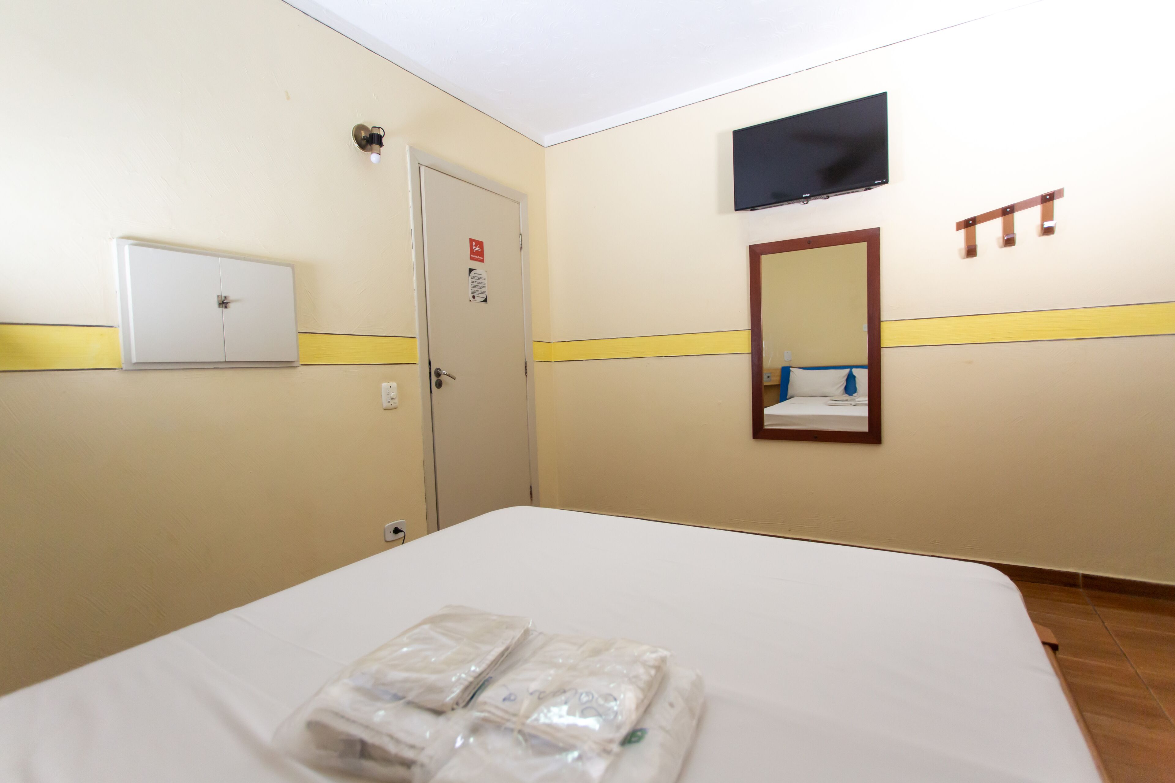 A hotel room with a bed, a door, a light switch, a wall-mounted TV, and a mirror.