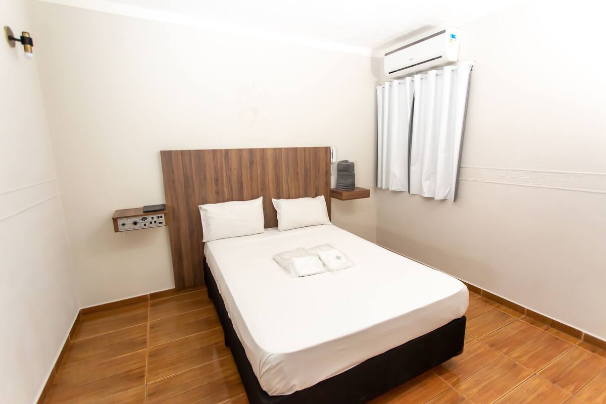 Een netjes opgemaakt bed met een houten hoofdbord, een witte muur en een klein plankje met een afstandsbediening.