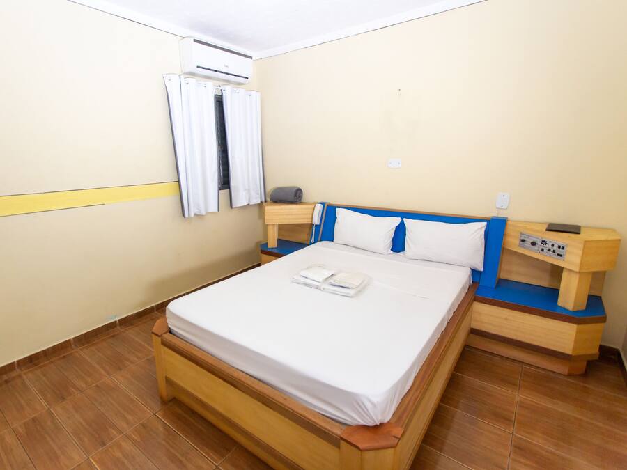 Een net opgemaakt bed met een witte matras en een houten hoofdbord. Er is een klein houten nachtkastje met een lamp en een stopcontact. De kamer heeft een betegelde vloer en een beige muur met een gele streep.