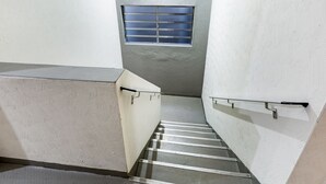 Una escalera estrecha con barandales metálicos, un pequeño ventanal con barrotes horizontales y una pared con textura.