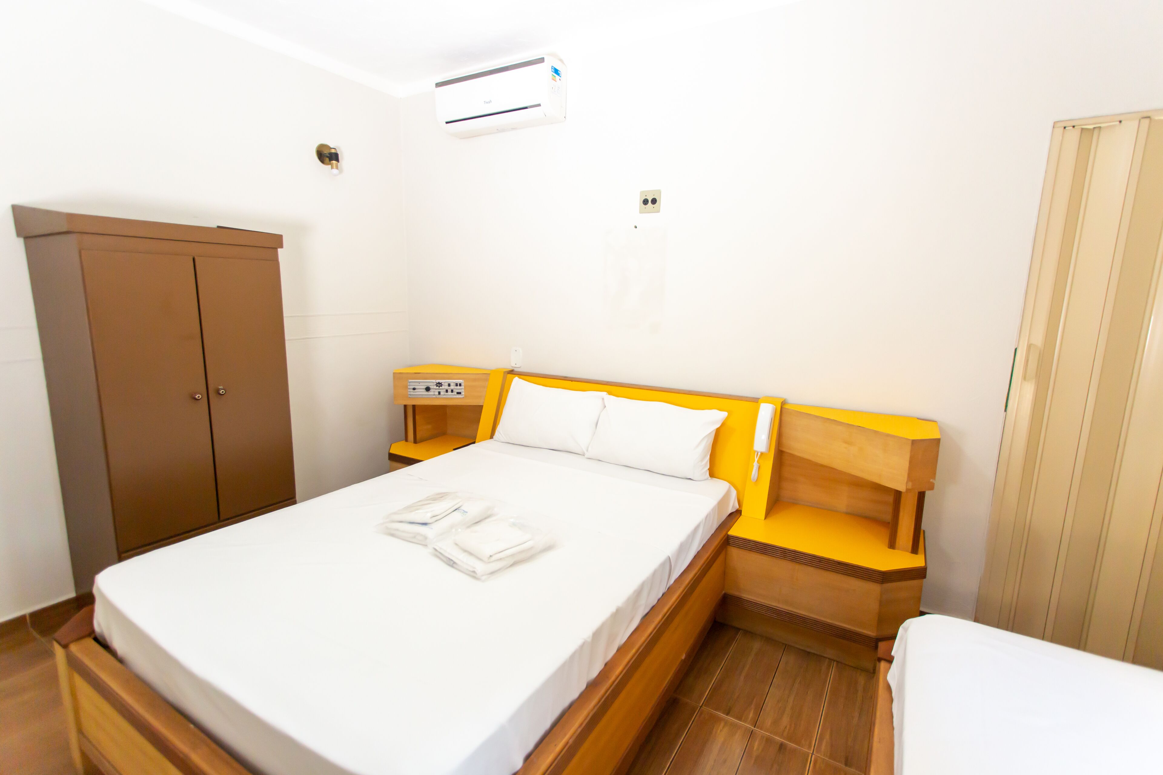 A hotel room with a bed, a wooden wardrobe, a bedside table with a lamp, and an air conditioning unit on the wall.